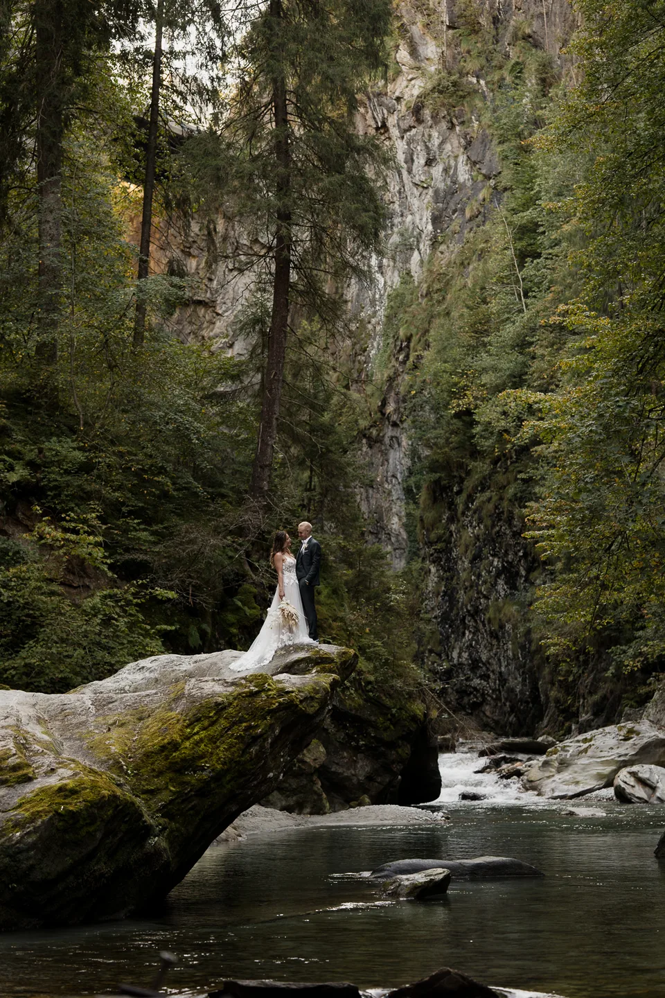 Braut im Brautkleid auf einem Felsen in einer feuchten, bewaldeten Schlucht am Wasser